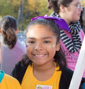 Girls on the Run girl smiling with purple ribbon in hair and glitter on face.
